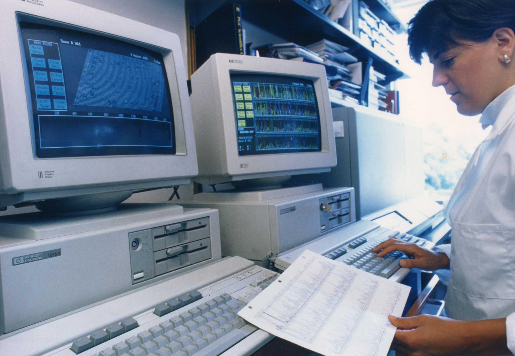 A researcher checks a computer readout from a sequencing machine while holding a Sanger sequencing chromatogram during the Human Genome Project.