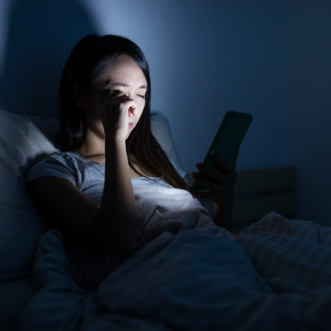 A stock photograph of a woman in bed looking at a phone in a dark room, to illustrate insomnia