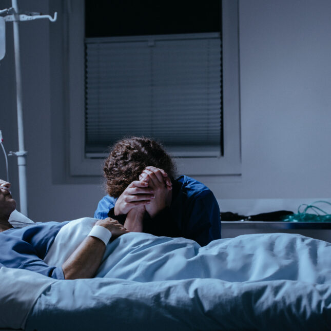 Next to a hospital bed, a person holds a patient's hand to their own face, as the patient receives treatment from an IV bag — health coverage from STAT