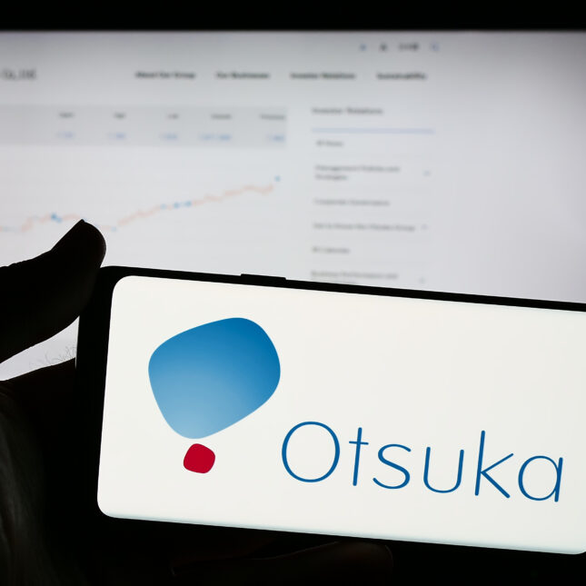 A hand holds a phone screen displaying the blue and red logo of Otsuka Pharmaceutical, in front of a laptop showing a stock chart — health tech coverage from STAT