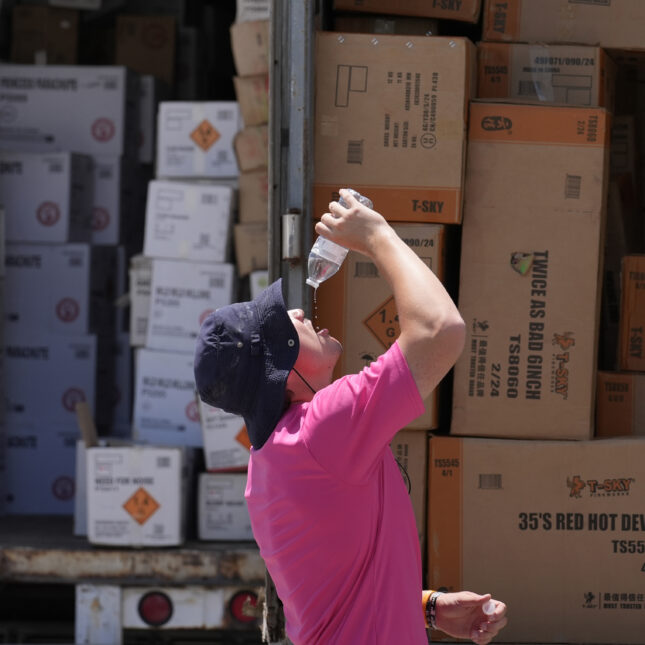 A person, wearing protective sun hat, tilts their head back to drink the last drop from a bottle outside a fireworks trailer — health coverage from STAT