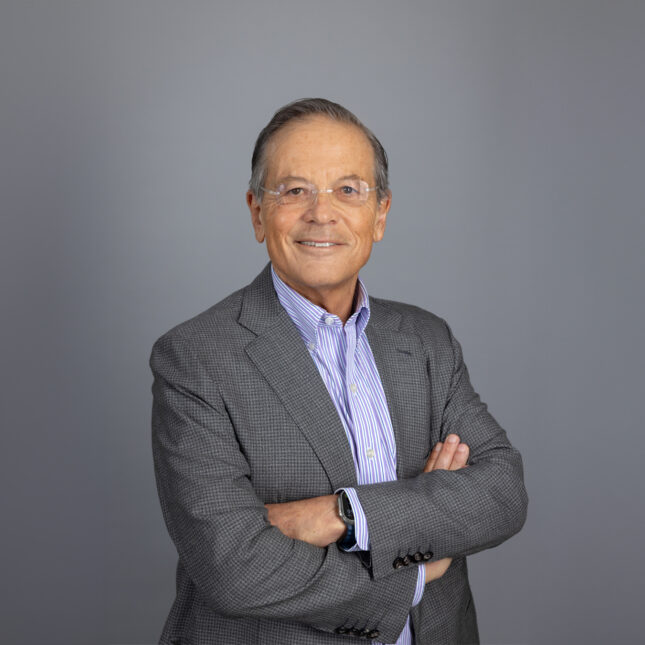 Arie Belldegrun, wearing glasses and a grey suit jacket, crosses arms as he stands for a portrait in front of a grey background.