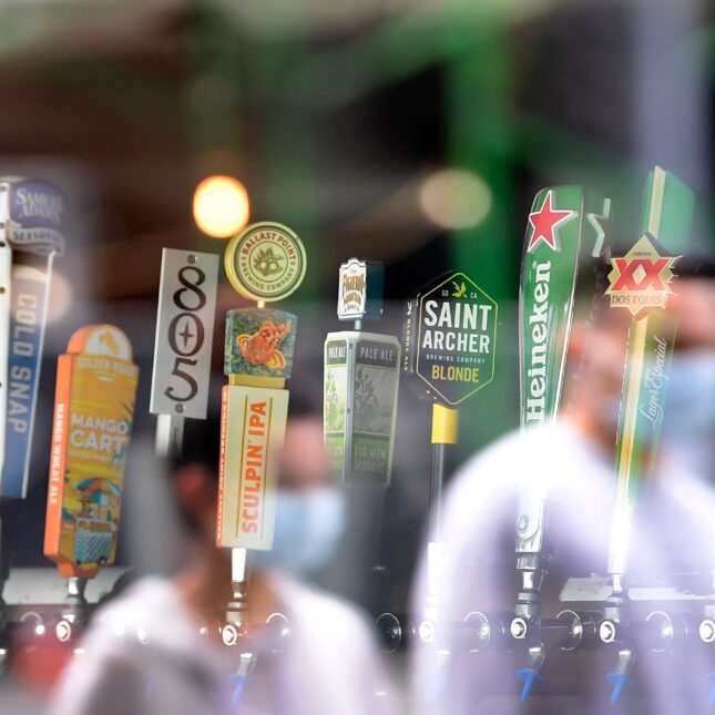 In daylight, a dozen beer taps are shown through a glass window, which also reflects two masked pedestrians — health coverage from STAT