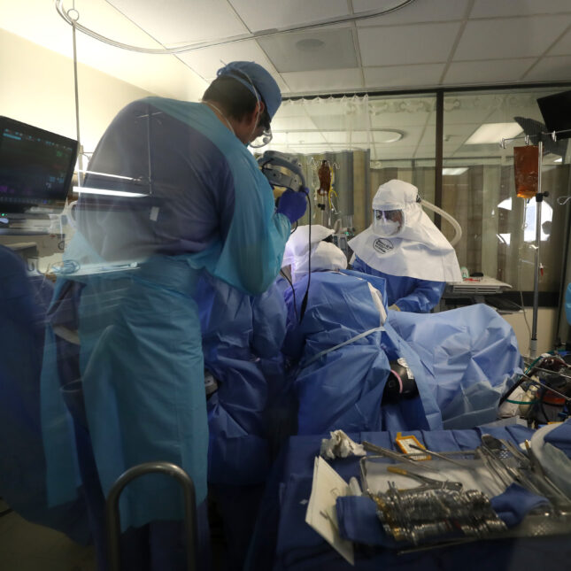 Inside an intensive care unit, a group of around six physicians wearing PPEs surround and perform a procedure on a COVID-19 patient — health coverage from STAT