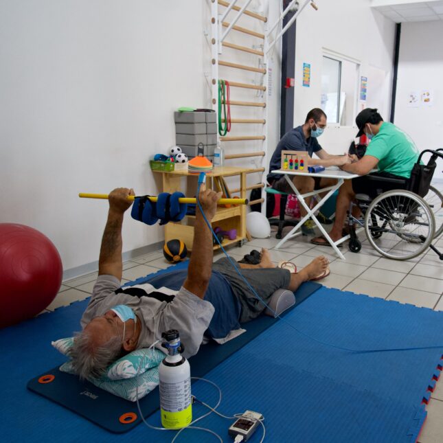 A masked person lays on floor mats, lifting weights while wearing a oximeter connected to a vital monitor. Two people work at a table in the background — health coverage from STAT