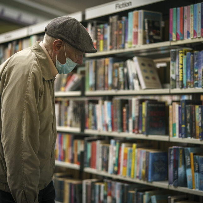 A photo of an older man wearing a wool cap and a surgical mask browsing the fiction stacks at a public library