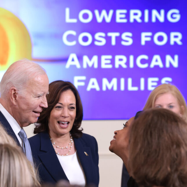 U.S. President Joe Biden and Vice President Kamala Harris converse with an audience in front of a projection screen that reads "LOWERING COSTS FOR AMERICAN FAMILIES" next to a pill bottle — politics coverage from STAT