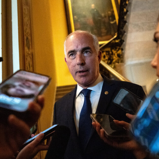 Sen. Bob Casey Jr. speaks in front of stairs while surrounded by a group of reporters as they record audio via phone — exclusive coverage from STAT