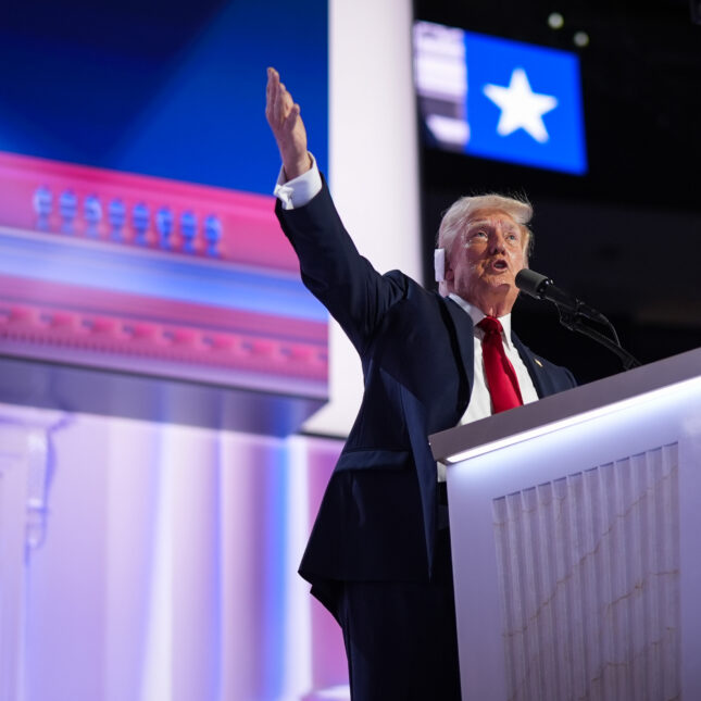 Republican presidential nominee, former U.S. President Donald Trump speaks at a podium in front of a projection screen, raising his right arm while wearing a ear bandage, a blue suit and a red tie — politics coverage from STAT