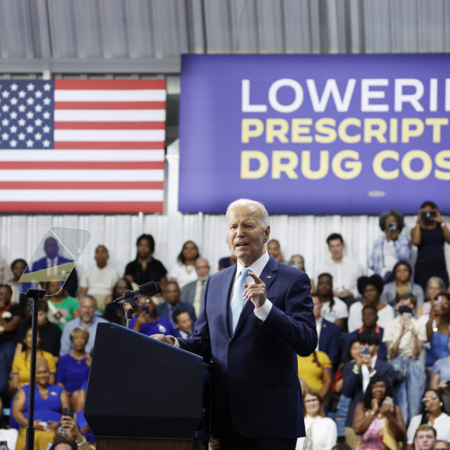 U.S. President Joe Biden scrunches his face while speaking at a podium. Behind him are audiences who sit and stand below a hung American flag on the left and a banner on the right that reads "LOWERING PRESCRIPTION DRUG COSTS" — politics coverage from STAT