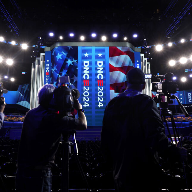 Four camera operators stand in front of the stage of the Democratic National Convention, where DNC 2024 and the national flag is projected — politics coverage from STAT