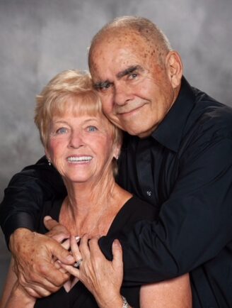 Frank Toro wraps his arms around his wife Bobbi Toro's shoulder, as Bobbi holds his arms. The couple posed in front of a gray backdrop — health coverage from STAT