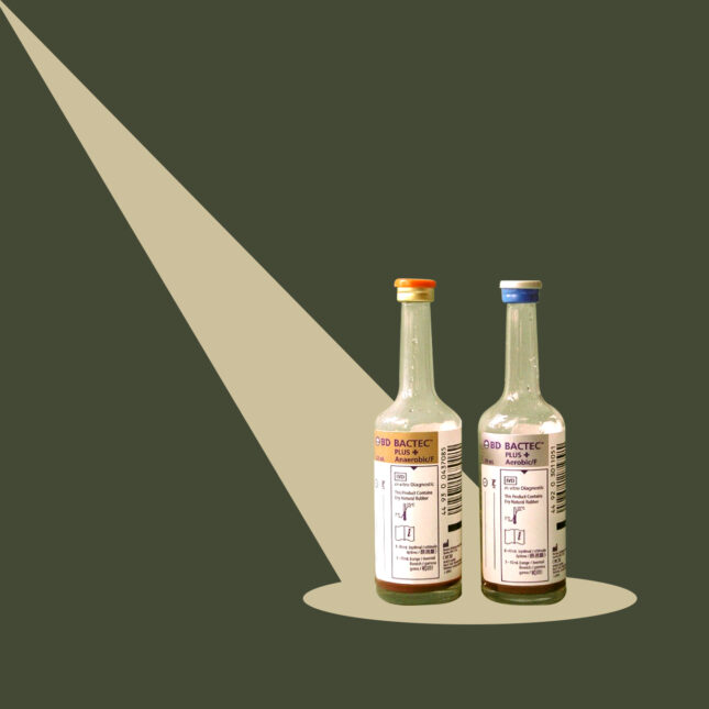 Two blood culture bottles stand together, spotlighted by khaki geometric shapes on a military green background — coverage from STAT